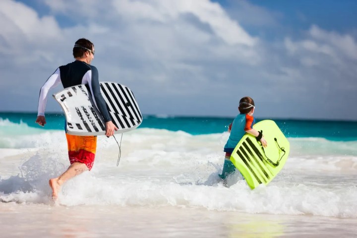 Two people with bodyboards running into the ocean on a sunny day.
