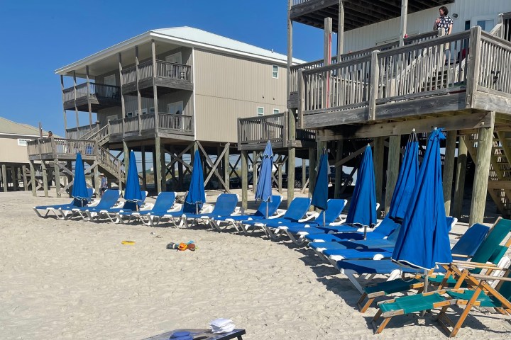 Row of beach chairs and umbrellas on sand near stilted beach houses.