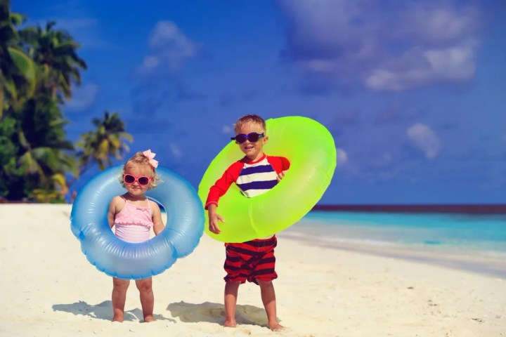Two children with inflatable rings on a sandy beach, wearing sunglasses.