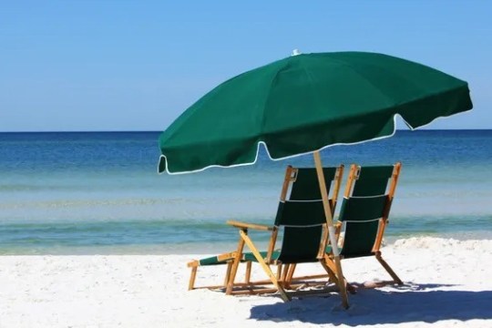 Two beach chairs under a large green umbrella on a sandy beach by the ocean.