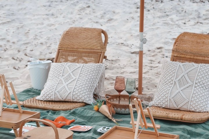 Beach setup with two chairs, cushions, umbrella, and picnic basket on a green-striped blanket.