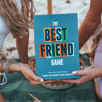 Two people holding 'The Best Friend Game' box on a picnic blanket.