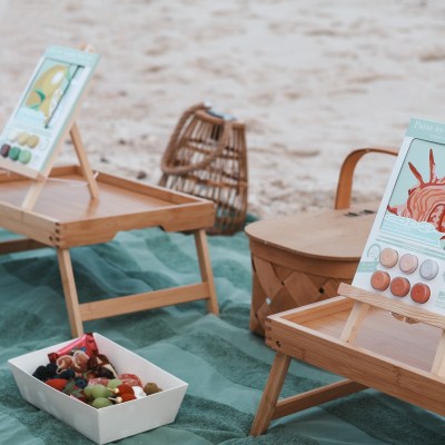 Beach picnic setup with trays, art, and snacks on a green blanket.