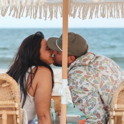 Couple kissing under a fringed umbrella on a beach.