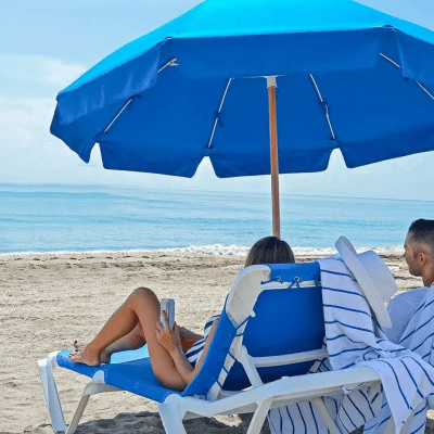Two people relaxing under a blue umbrella on beach chairs by the sea.