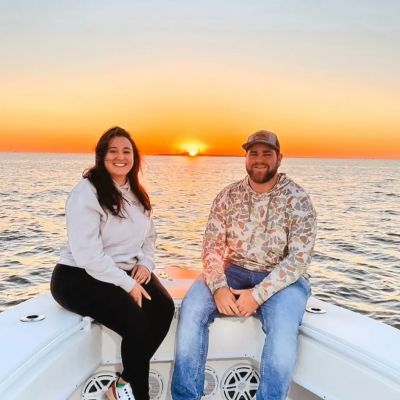 Two people sit on a boat's bow at sunset, smiling at the camera.